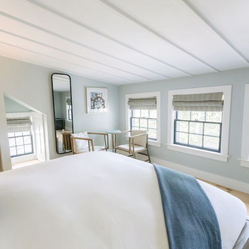 Bright bedroom with a bed, two armchairs, large windows with blinds, a wall mirror, and framed art under a sloped ceiling.