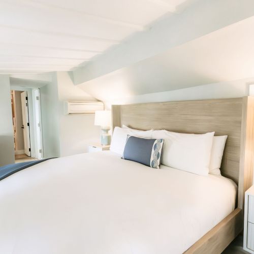 A bright modern bedroom with a large bed, pillows, bedside lamps, a dresser, TV, and a wall-mounted air conditioner.