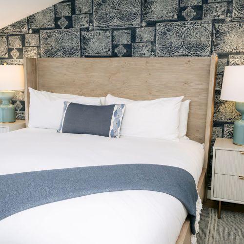 A cozy bedroom with a wooden bed, white linens, two bedside lamps, and a dark vintage world-map wallpaper behind it.