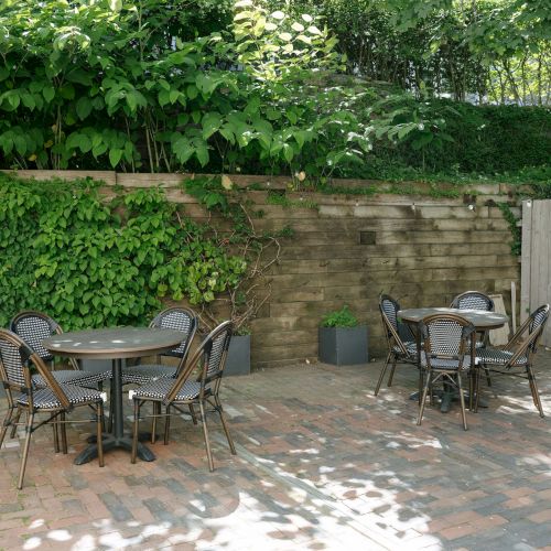 A green, ivy-covered backyard patio with two metal tables and chairs, hanging string lights, and a wooden fence along the wall.