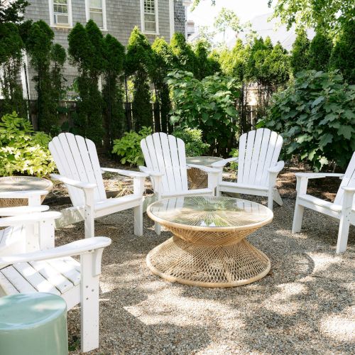 White Adirondack chairs circle a wicker patio table in a lush garden courtyard with greenery and a house wall behind.