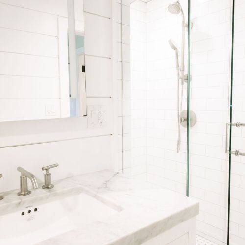 A bright white bathroom with a marble sink, chrome faucets, and a clear glass shower enclosure with handheld showerhead.
