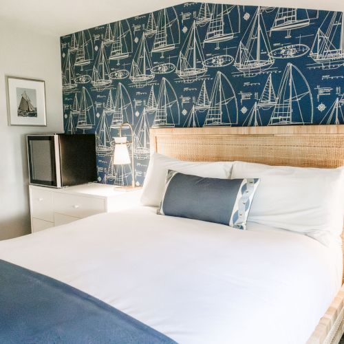 A bright hotel bedroom with a large bed, nautical sailboat wallpaper, bedside lamps, and white dresser with mirror.