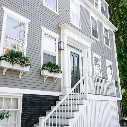 A multi-story white house with white trim, green steps, windows, a porch railing, and hanging planters by the entrance.