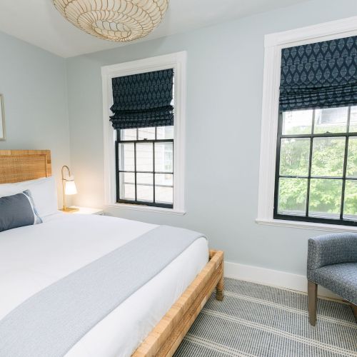 Bright bedroom with a bed, blue patterned shade blinds on two windows, a sitting chair, and a woven ceiling pendant.