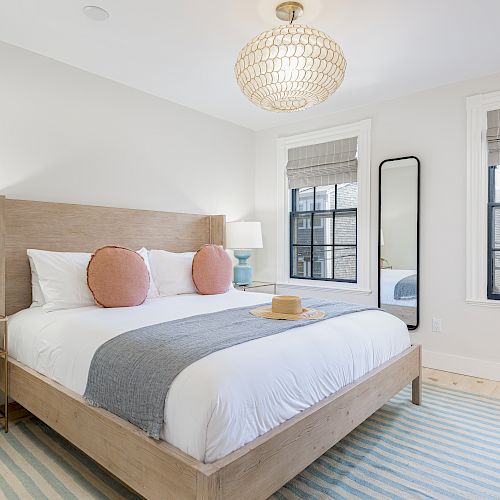 A modern bedroom with a large bed, wooden frame, and two coral pillows. There are side tables with lamps, a full-length mirror, and a chandelier.