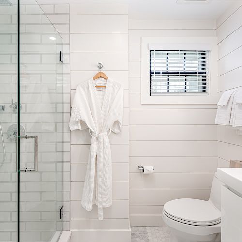 This image shows a modern bathroom with a glass shower, a white robe hanging, towels on a rod, a toilet, and a window with blinds.