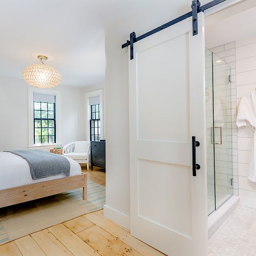 A modern bedroom with wooden flooring, a bed with white and grey sheets, a sliding barn door leading to a bathroom with a robe hanging on the wall.