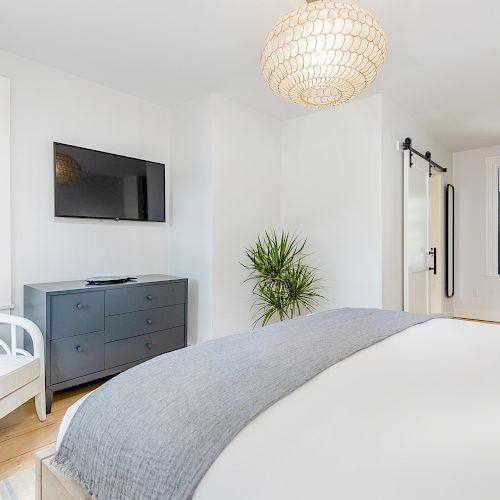 A bright, modern bedroom with a large bed, a wall-mounted TV, a dresser, a chair, a potted plant, and large windows.