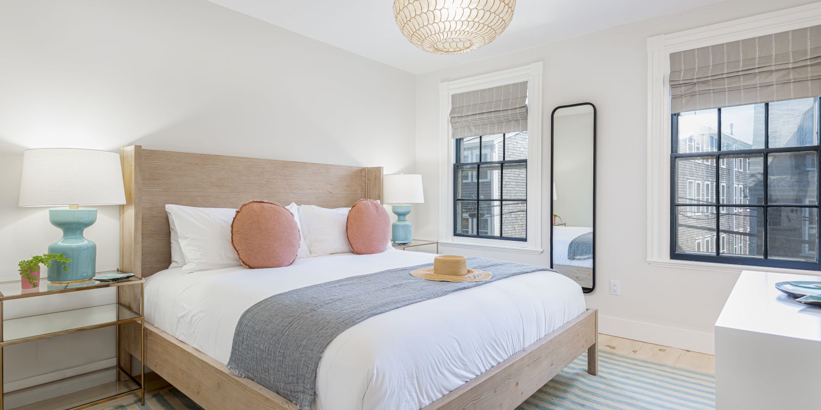 A cozy bedroom with a large bed, two pink pillows, a gray throw, bedside tables with lamps, a mirror, and windows letting in natural light.