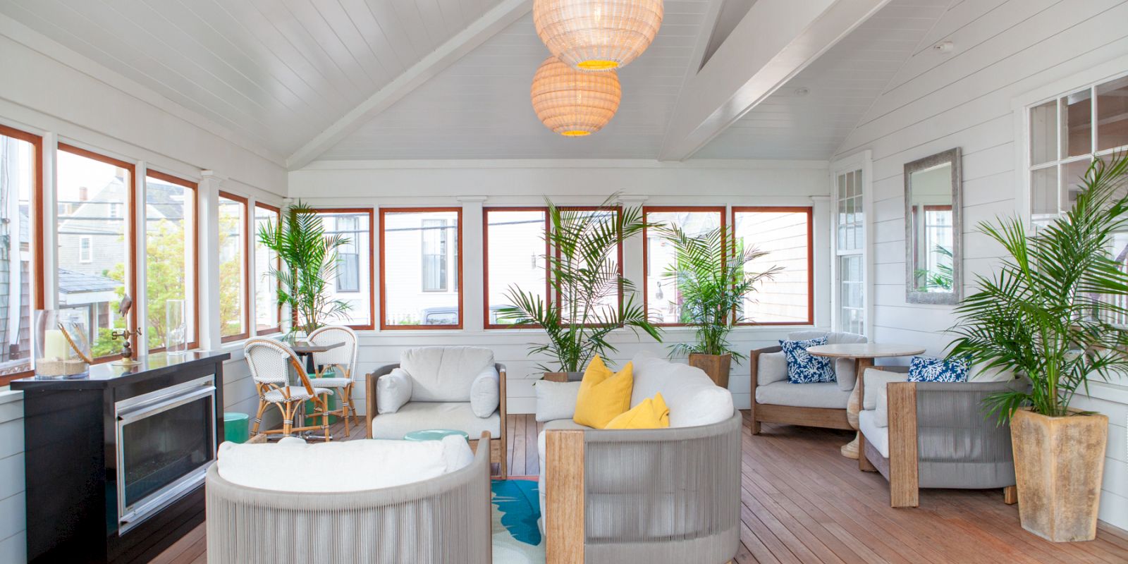 A bright, modern sunroom with large windows, comfortable furniture, indoor plants, and a fireplace, featuring wooden floors and hanging lights.