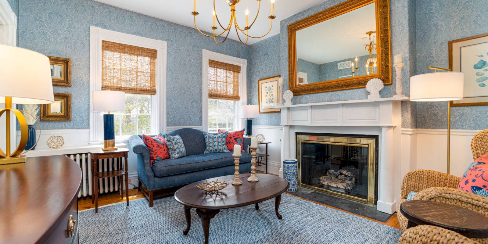 A cozy living room with blue wallpaper features a chandelier, a fireplace, a large mirror, a navy sofa, wicker chairs, and wooden furniture.