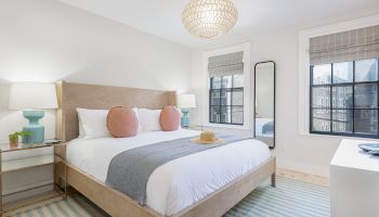 A modern bedroom with a neatly made bed, two side tables, lamps, a tall mirror, two windows, and a light fixture above, all in a bright, airy space.