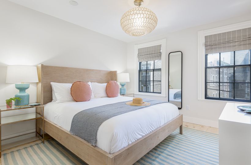 A modern bedroom with a neatly made bed, two side tables, lamps, a tall mirror, two windows, and a light fixture above, all in a bright, airy space.