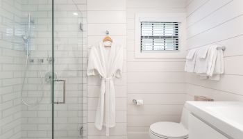 The image shows a clean, modern bathroom with a glass shower, a hanging white robe, a toilet, a sink, towels on a rail, and a small window with blinds.