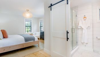 A cozy bedroom with a bed, chandelier, sliding barn door leading to a bathroom with a glass shower and a bathrobe hanging.