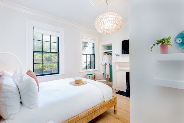 A bright, cozy bedroom features a white bed, wooden accents, large windows, a hanging light, indoor plant, and a hat resting on the bed.