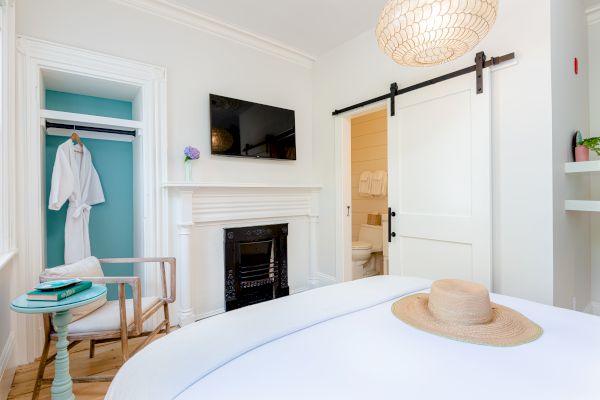 A cozy bedroom with a bed, a straw hat, a small table, a chair, a TV, a fireplace, a hanging robe in the closet, and a sliding door to a bathroom.