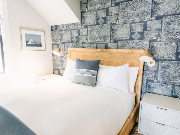 A bright bedroom with a large bed, white linens, wicker headboard, bedside lamps, and blue nautical world-map wallpaper.