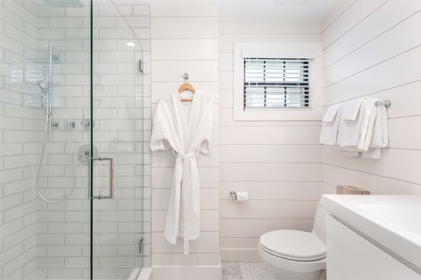 A modern bathroom with a glass shower, a robe hanging, a window, white tiles, towels on a rack, a toilet, and a countertop with a sink.