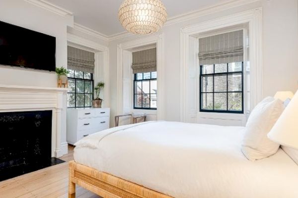 A bright bedroom features a bed with white linens, a black fireplace, a wall-mounted TV, a white dresser, plants, and two large windows with blinds.