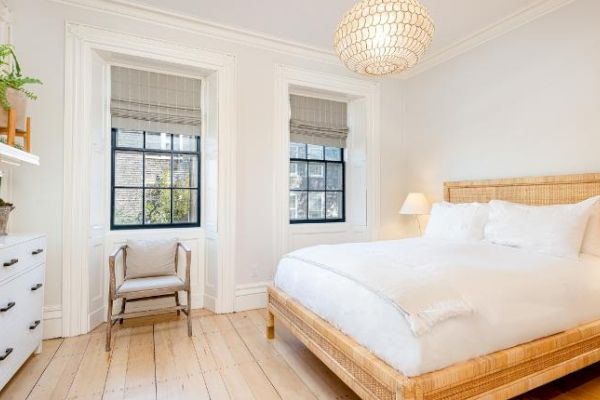 A bright bedroom with a wicker bed frame, white bedding, a chest of drawers with plants, a chair, and two windows with blinds, and a hanging lamp.