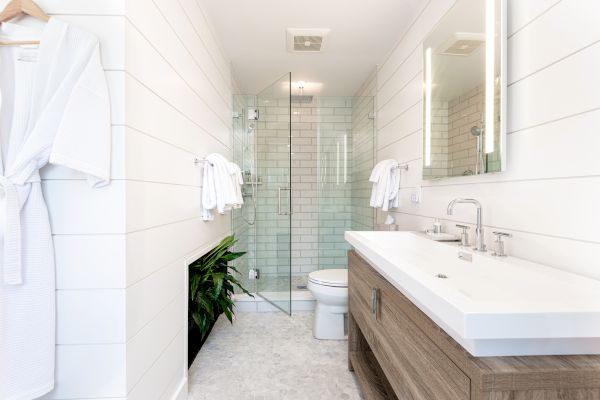 A modern bathroom with a wooden vanity, white countertop, large mirror, glass shower, toilet, wall-mounted towel racks, and a robe hanging on the wall.