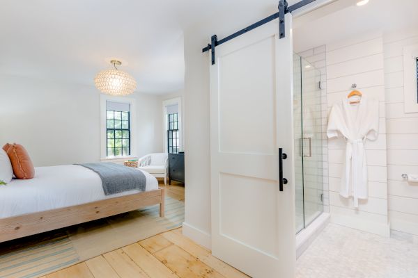 A cozy bedroom with a bed, a chandelier, and two windows, next to a bathroom with a sliding door, featuring a shower and a hanging robe.