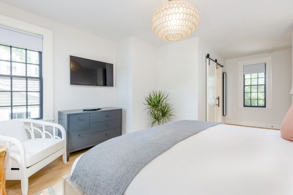 A modern bedroom with a cozy bed, wall-mounted TV, dresser, lounge chair, and plant, featuring large windows and a sliding barn door.