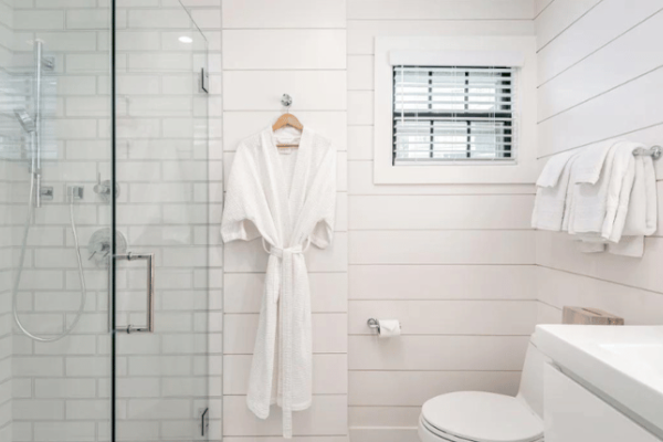 The image shows a bright, modern bathroom with a glass shower enclosure, a white bathrobe hanging, towels on a rack, and a toilet underneath a window.