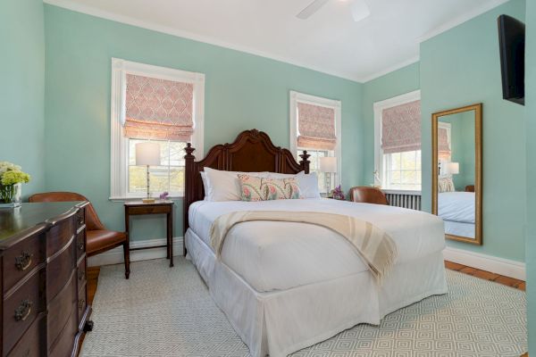 A neatly arranged bedroom with a double bed, wooden furniture, green walls, and a large mirror. Windows have fabric blinds.