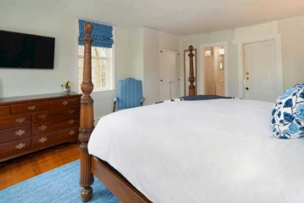 A bedroom with a large bed, wooden furniture, TV on the wall, blue accents, and windows. White walls and a blue rug complete the decor.