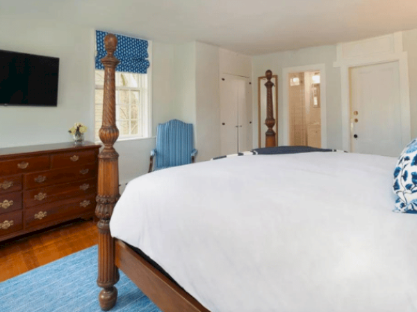 A bedroom with a large bed, wooden furniture, TV on the wall, blue accents, and windows. White walls and a blue rug complete the decor.