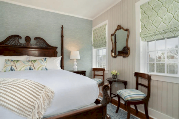 A cozy bedroom with a large bed, wooden headboard, side table with lamp, two chairs, small table, antique mirror, and two windows with patterned blinds.