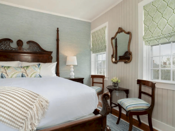 A cozy bedroom with a large bed, wooden headboard, side table with lamp, two chairs, small table, antique mirror, and two windows with patterned blinds.