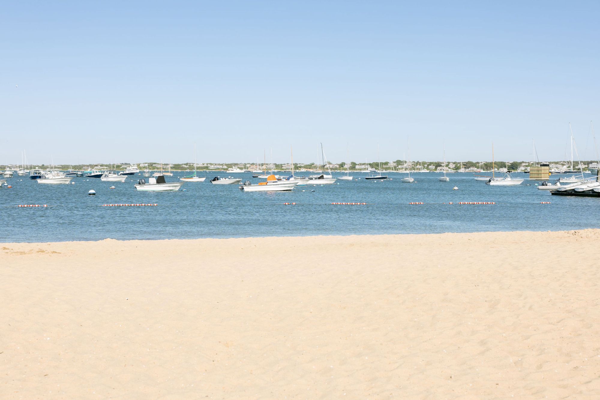 A calm sandy beach overlooks a marina filled with sailboats and yachts moored in blue water under a clear sky.