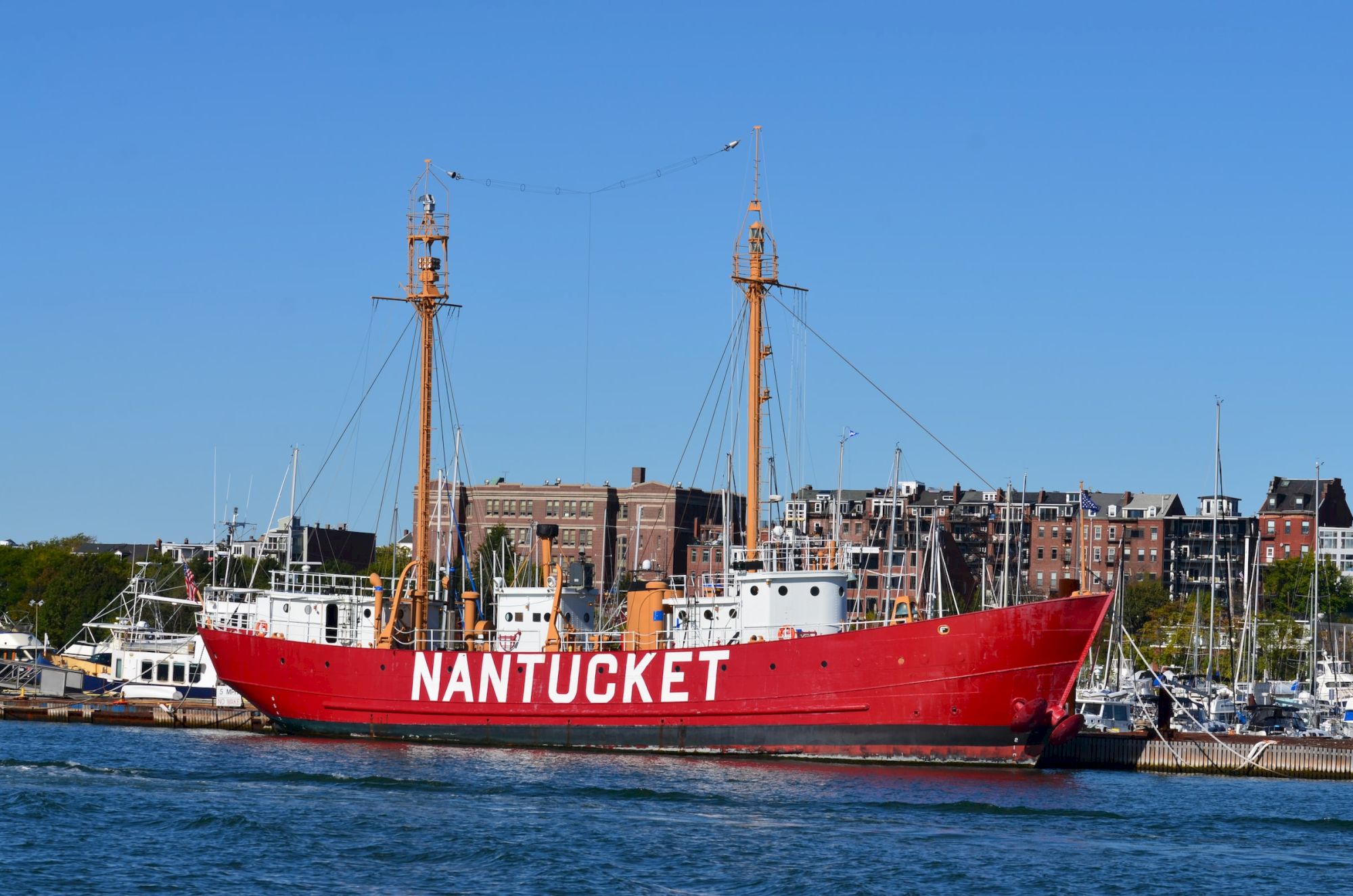 A red ship labeled "NANTUCKET" is docked in a harbor, with buildings and other boats in the background, under a clear blue sky.