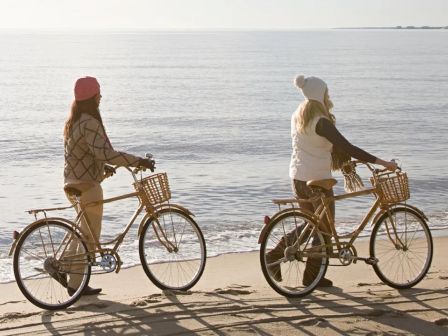 Two people wearing winter attire ride bicycles along a serene beach with calm ocean waves in the background.