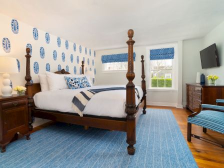 A bedroom with a four-poster bed, blue-themed decor, a dresser with a TV, a chair, and a window with printed blinds.