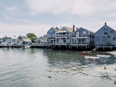 This image shows a waterfront scene with several gray wooden buildings on stilts, small boats tied up, and calm water reflecting the structures.