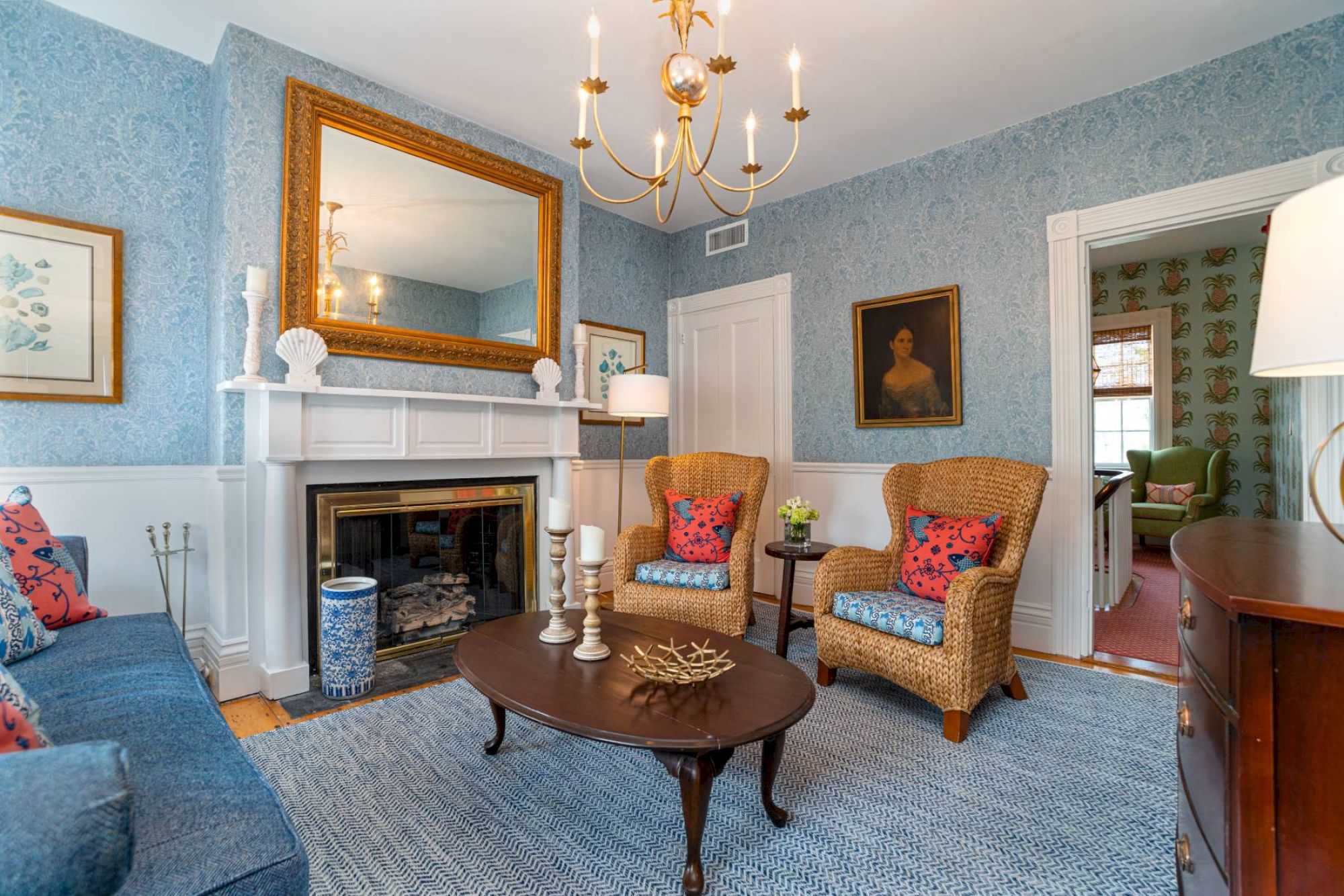 This image shows a cozy living room with patterned wallpaper, a large mirror above a fireplace, wicker chairs, and a wooden coffee table.