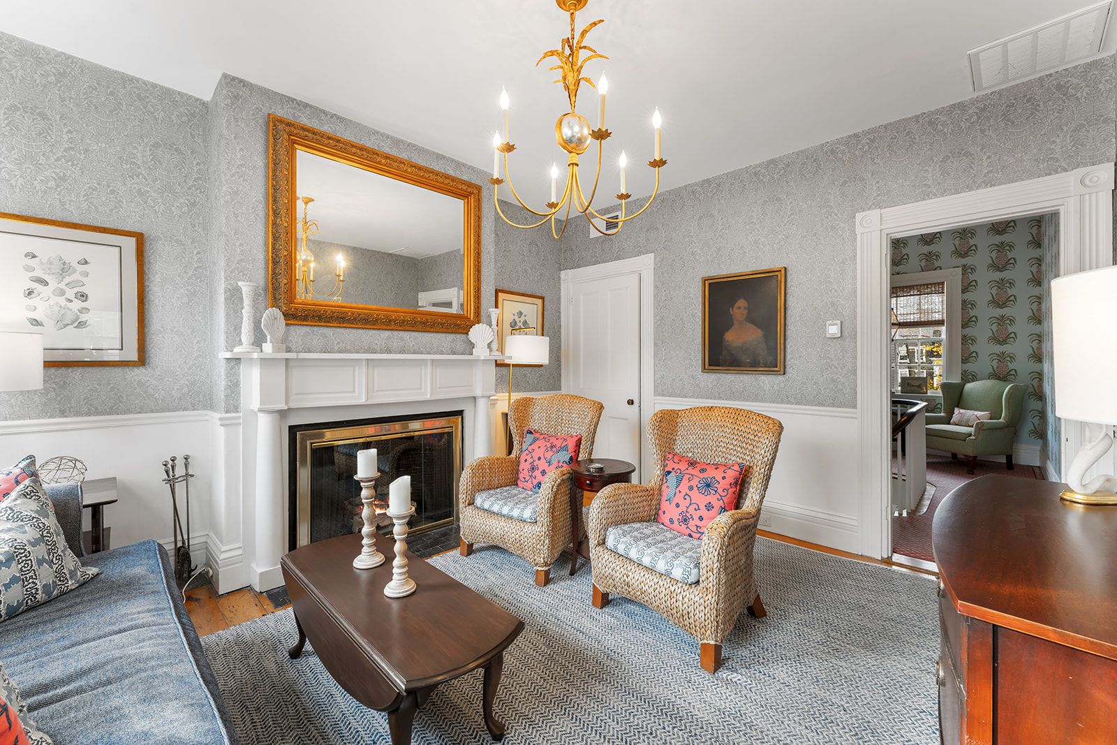 A cozy living room with a chandelier, fireplace, armchairs, and a mirror above the mantle, styled in classic decor with neutral tones.