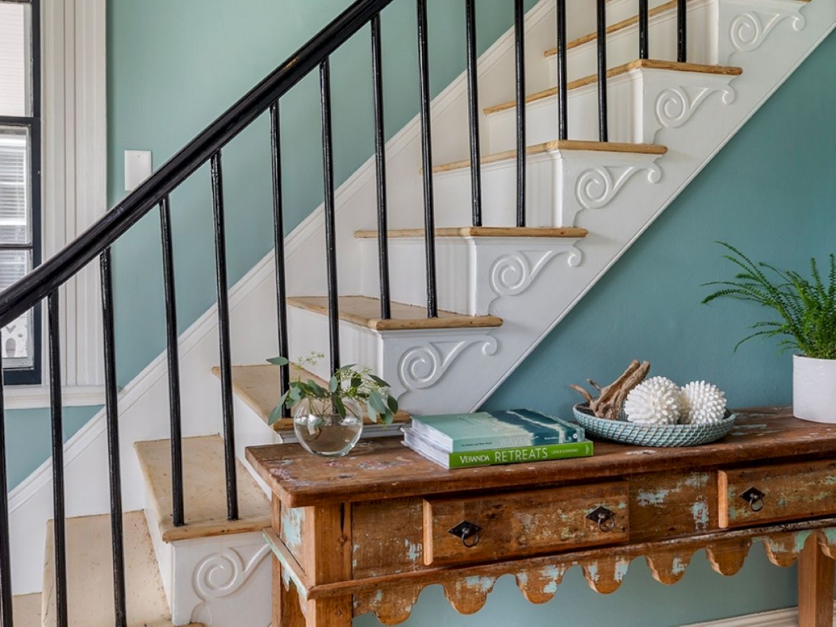 A rustic wooden table sits by a staircase with vases, books, and decorative items. The walls are painted teal, and a window is on the left side.