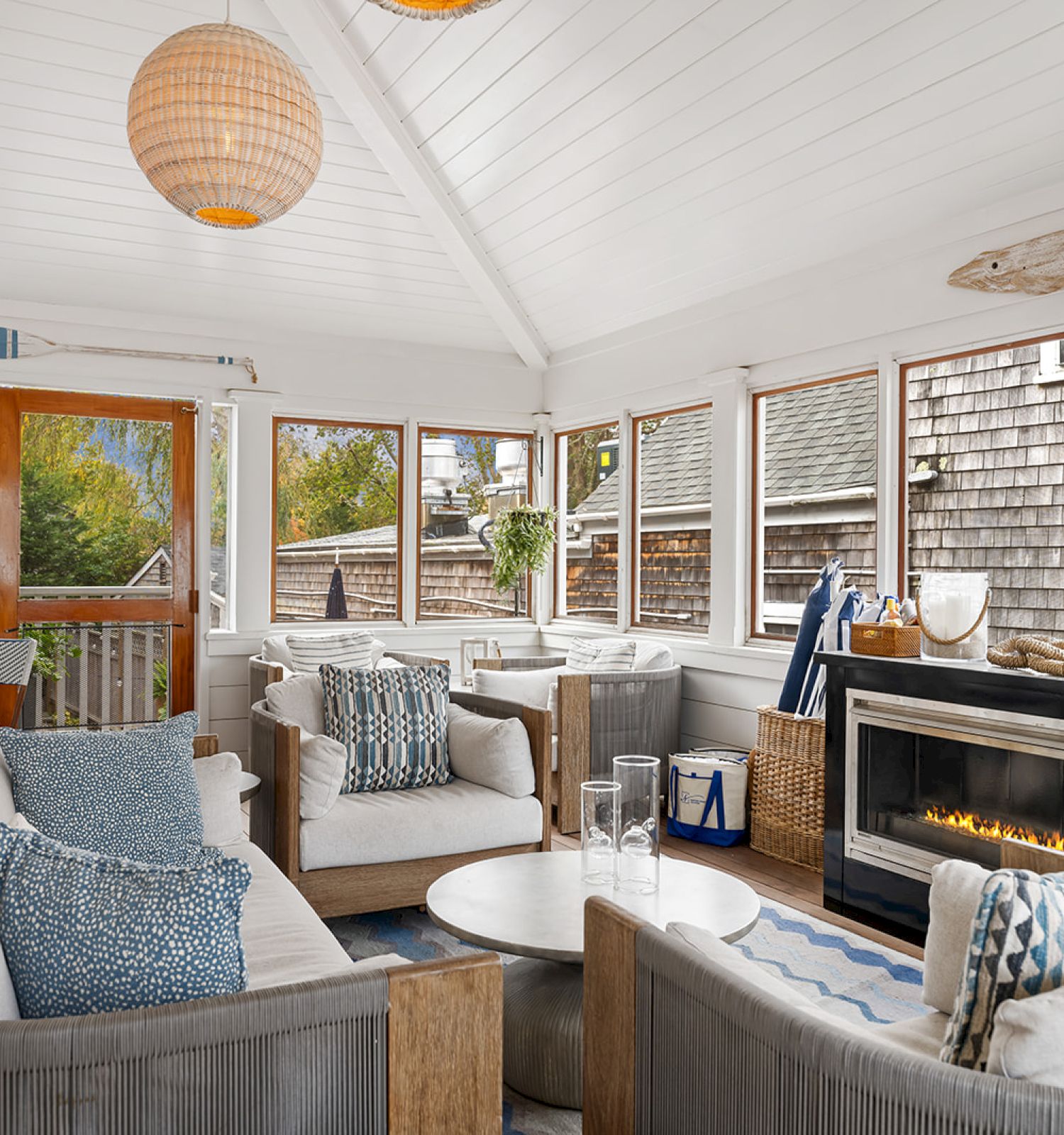 A cozy sunroom with wicker furniture, blue cushions, a wooden ceiling, a fireplace, and nautical decor like an oar and fish wall art.