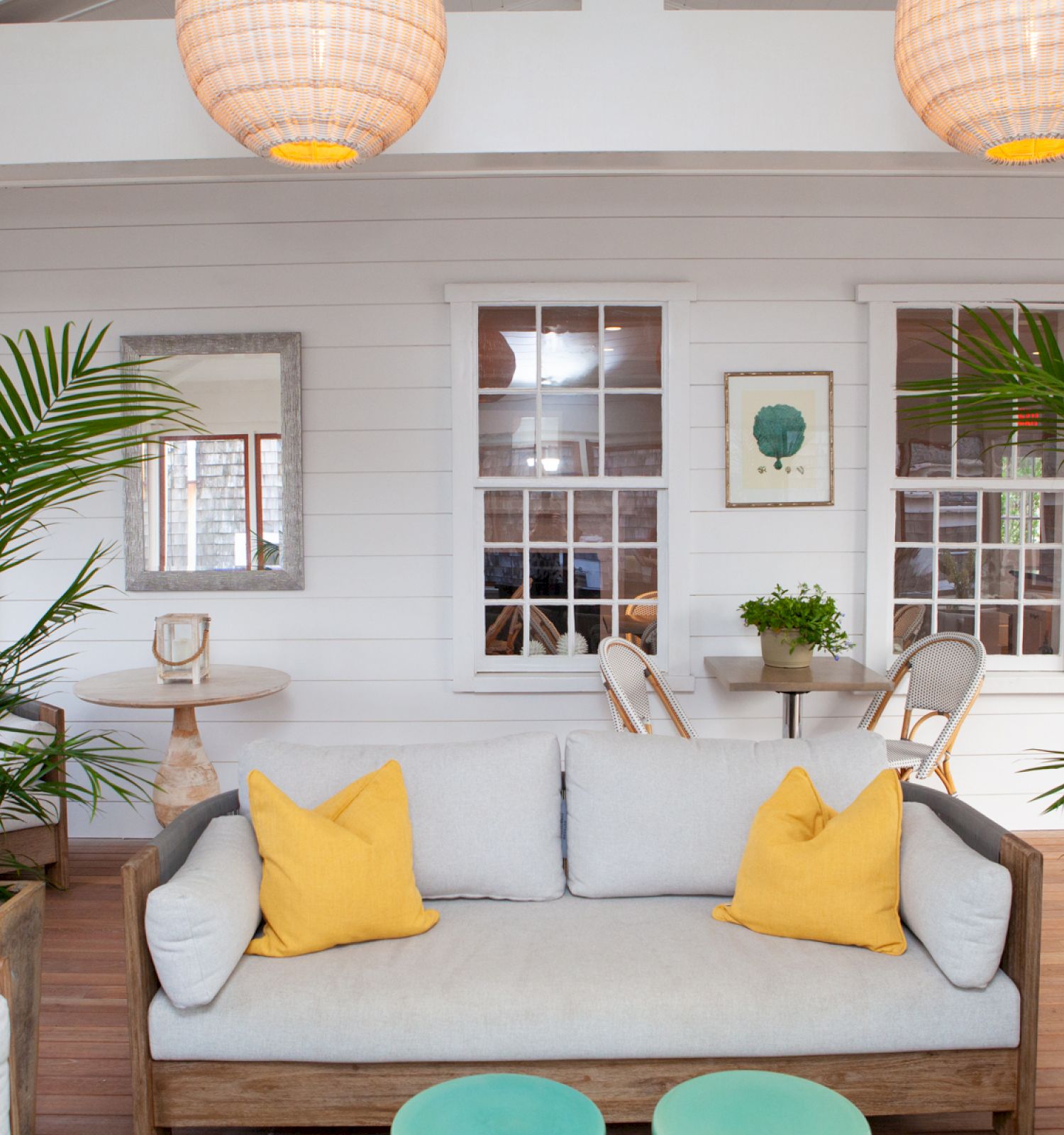 A cozy indoor space with a wooden couch, yellow pillows, potted plants, hanging lights, and a round table surrounded by chairs.