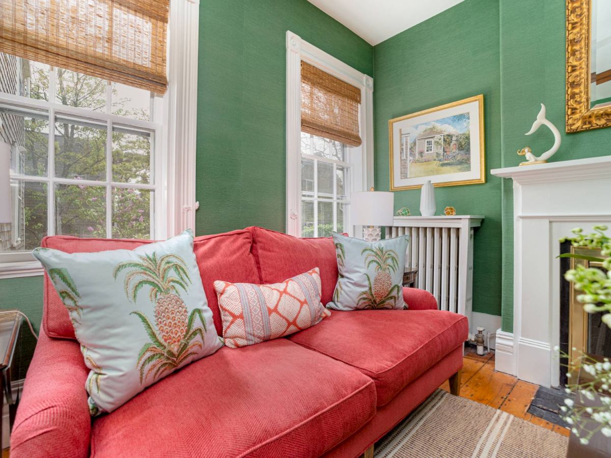 A cozy living room with green walls, a red couch with pineapple-themed pillows, large windows, a fireplace, and wall art.