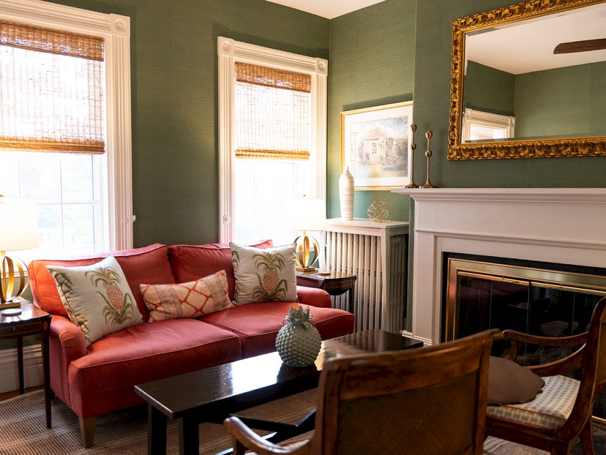 A cozy living room with green walls, red sofa, decorative cushions, fireplace, and wooden furniture, featuring a gold-framed mirror.
