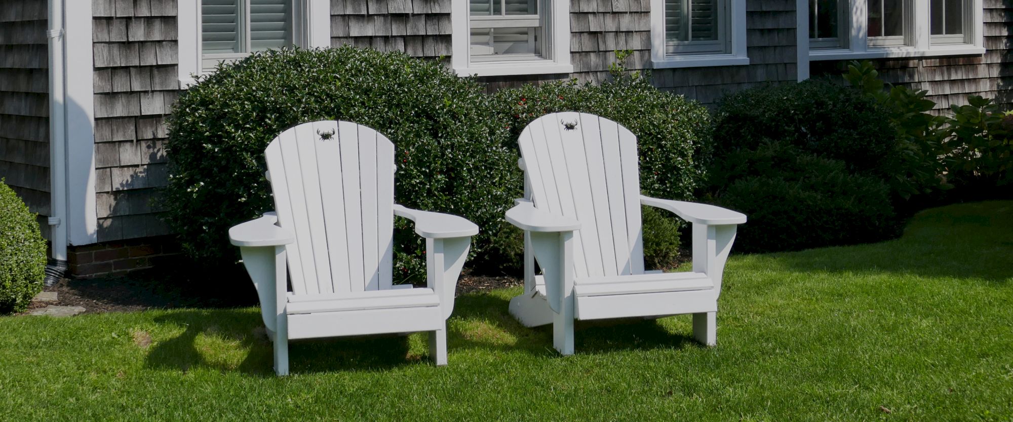 The image shows two white Adirondack chairs on a well-manicured lawn in front of a house with shingled siding and green bushes.