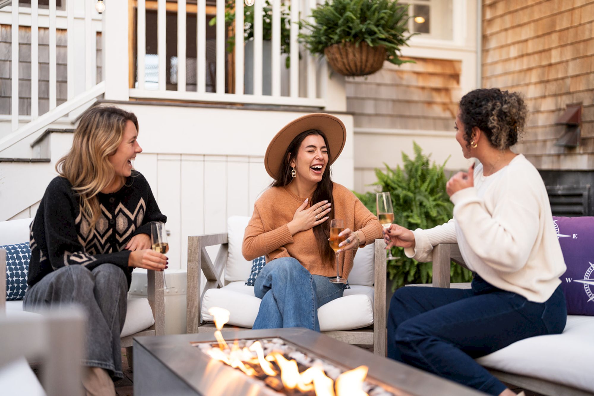 Three people are seated outdoors, enjoying drinks and laughing around a fire pit.