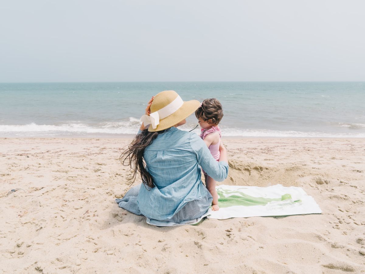 A woman with a hat sits on the beach, holding a child, both facing the ocean on a sunny day, with a towel beneath them in the sand.